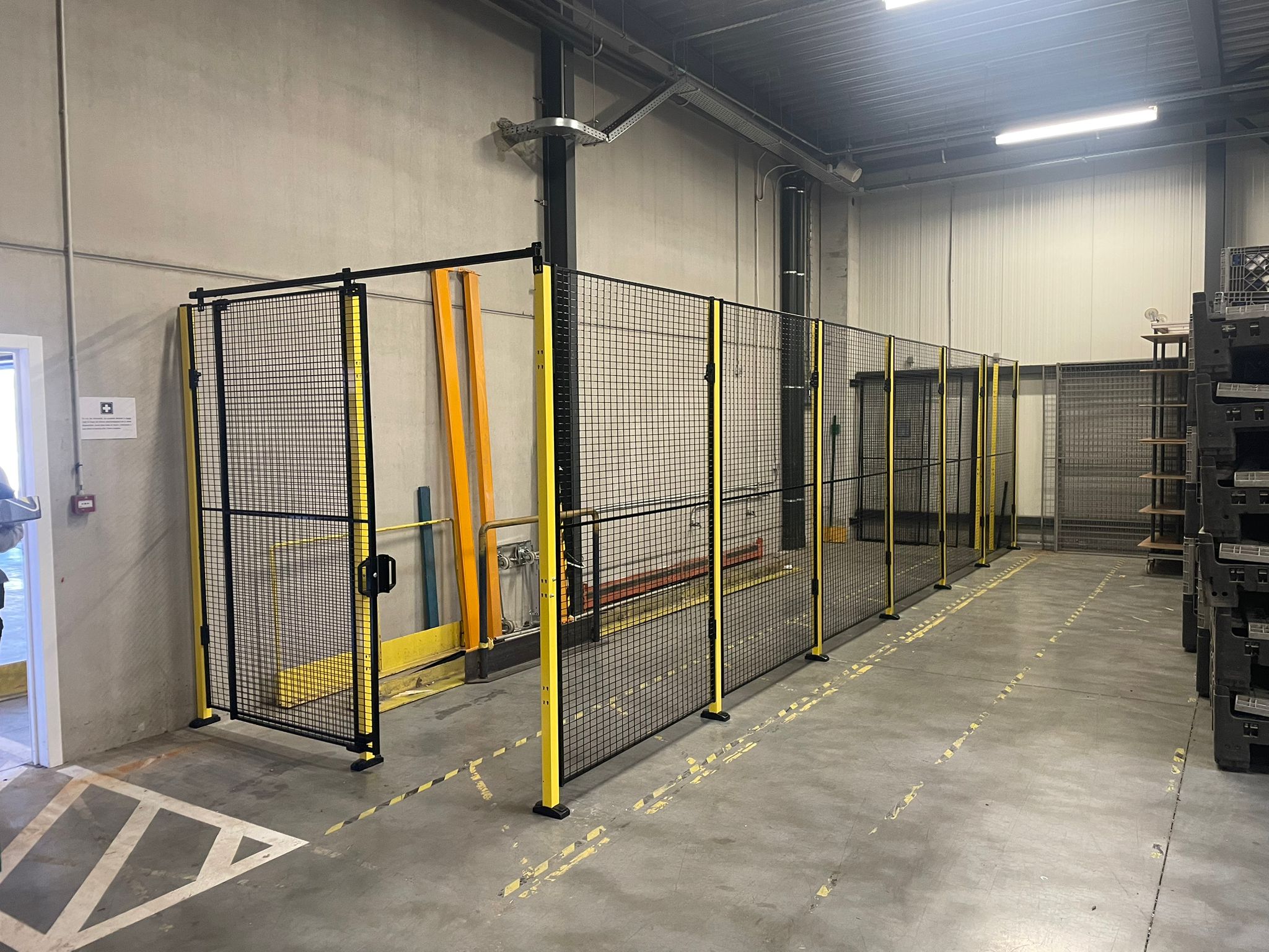 Lang hekwerk met zwart gaaswerk en gele staanders langs een hefinstallatie in een magazijnruimte, met geel-zwarte vloermarkeringstape en een hijsinstallatie aan het plafond