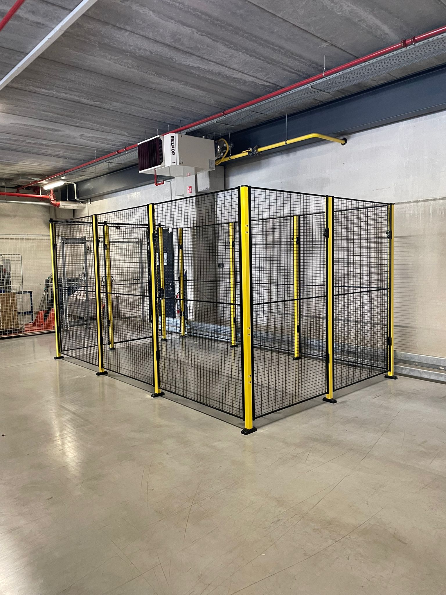 Hekwerkruimte met zwart gaaswerk en gele staanders rond een technische installatie in een magazijn, met een Reznor verwarmingstoestel aan het plafond