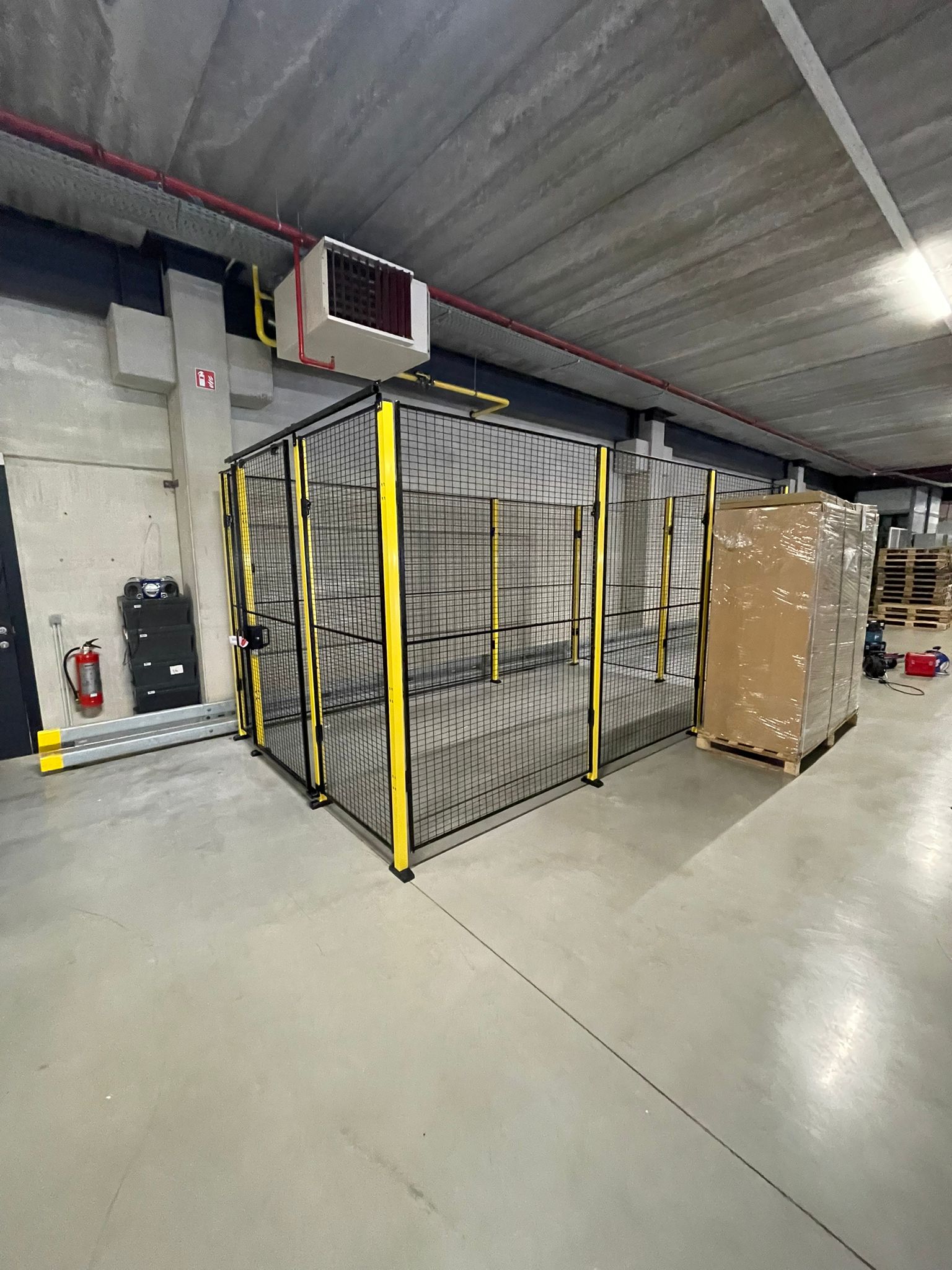 Gesloten hekwerkruimte met zwart gaaswerk en gele staanders in een ondergronds magazijn van een printdistributiebedrijf, met een brandblusser en elektrisch schakelkast op de achtergrond