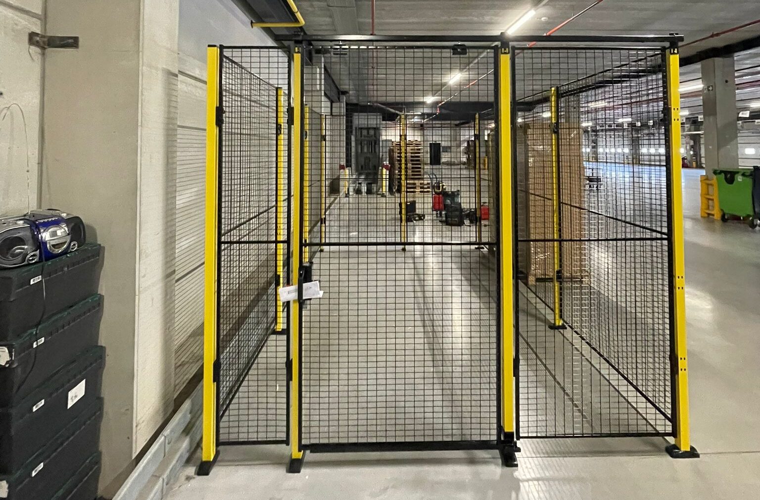 Smal doorgangshek met zwart gaaswerk en gele staanders als toegangspoort tot een lange ondergrondse gang in een distributiecentrum