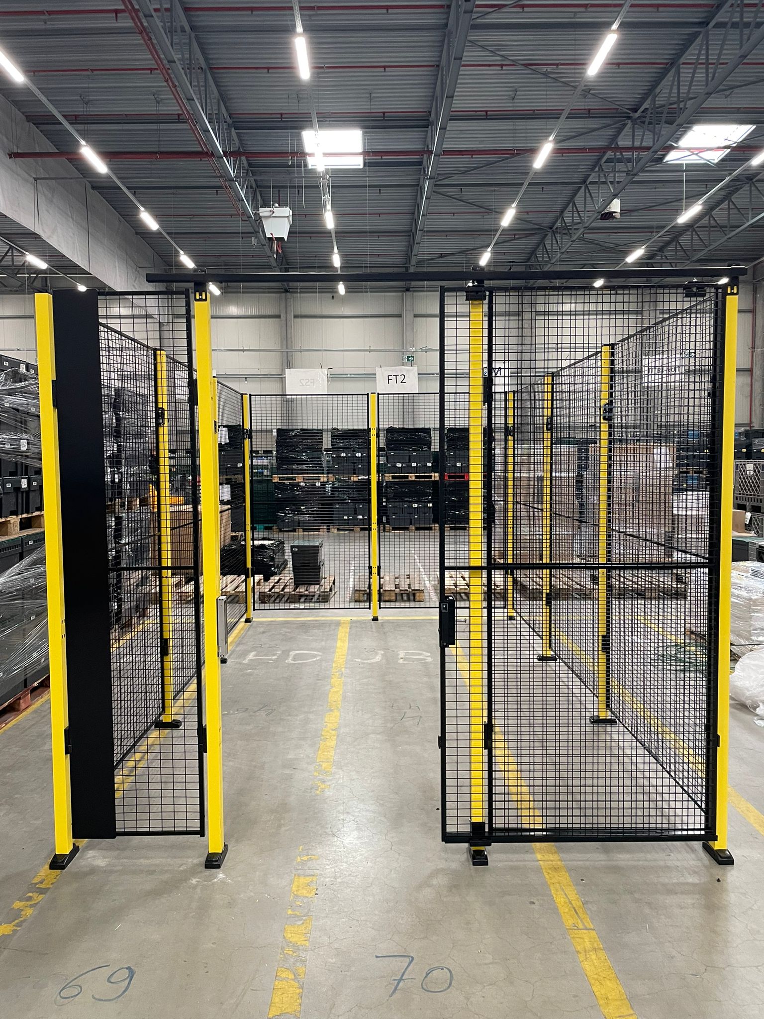 Zwart gaaswerk hekwerk met gele staanders als doorgangspoort in een actief magazijn met genummerde vakken, pallets en magazijnstellingen op de achtergrond