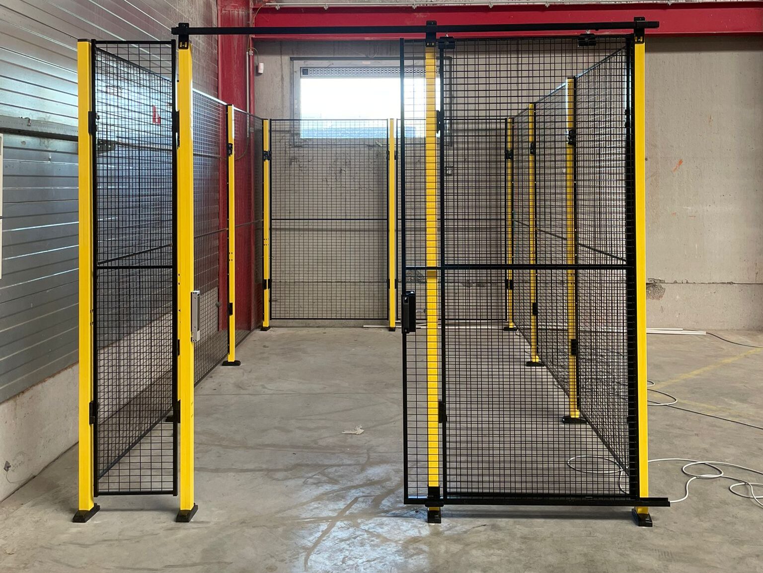 Doorkijkopname door een open hekwerkdoorgang met zwart gaaswerk en gele staanders naar een lege industriële ruimte met een laadkaaideurdeur op de achtergrond