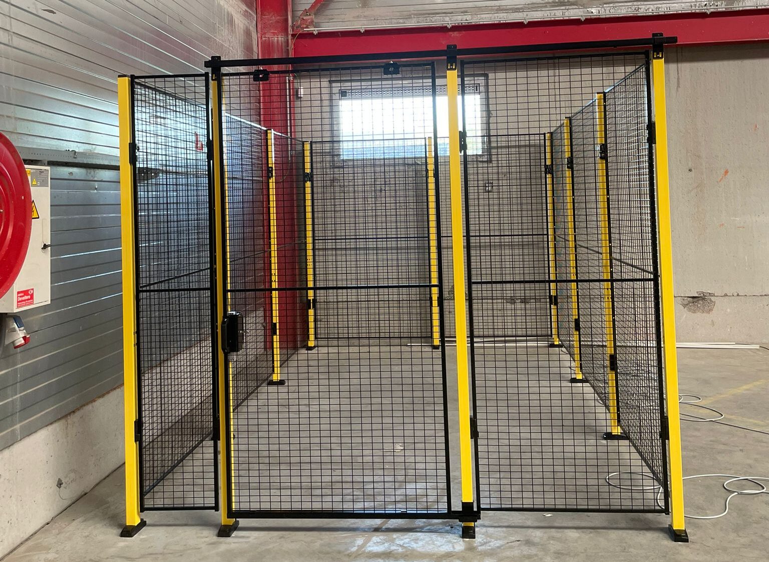 Frontaanzicht van een breed zwart hekwerk met gele staanders en twee ingangszones in een lege industriële ruimte met rode staalconstructie