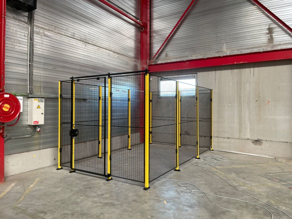 Zwart hekwerk met gele staanders en twee afzonderlijke toegangsdeuren in een industriële hal met rode staalconstructie en brandslangkast op de achtergrond