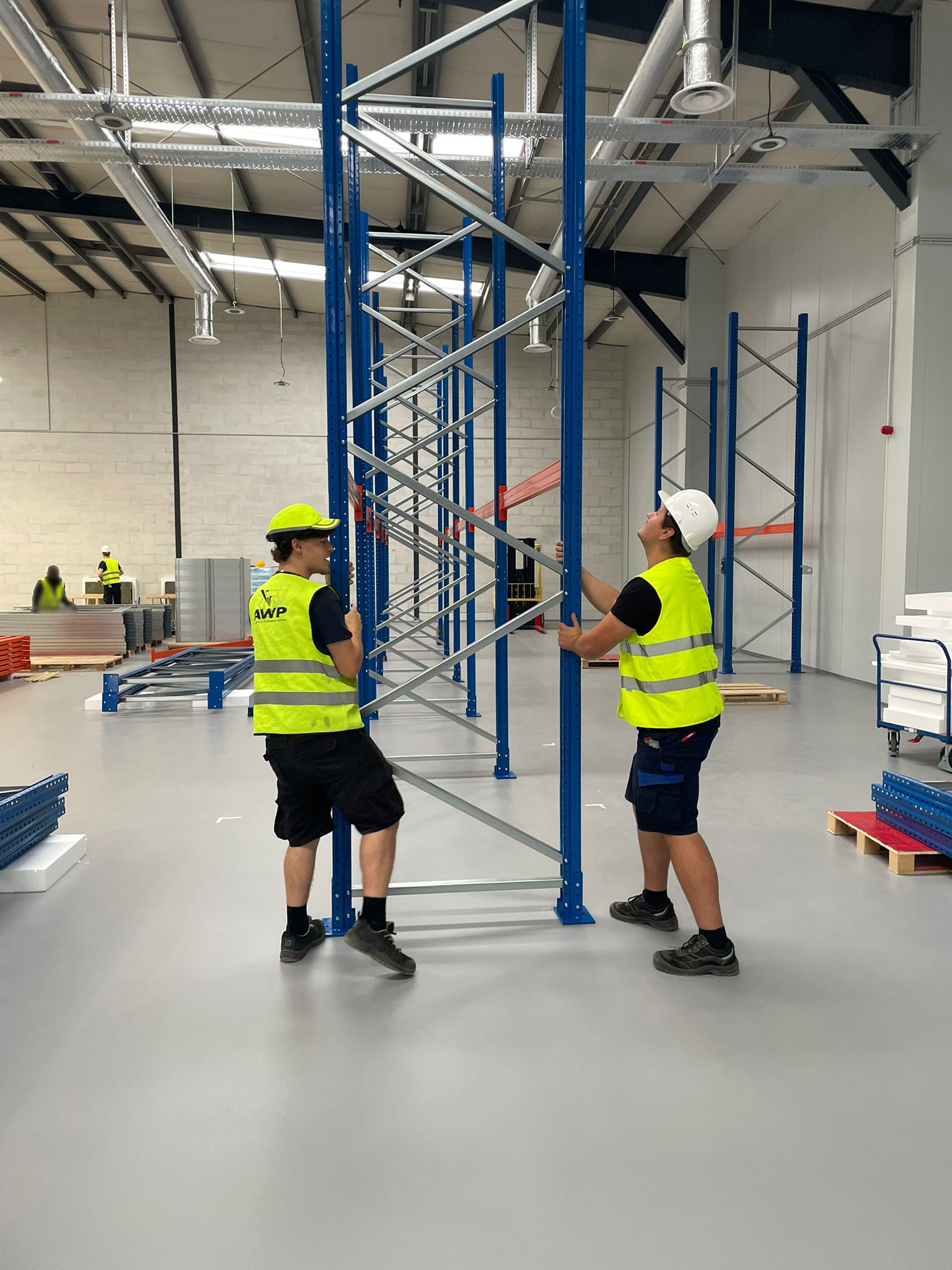 Twee AWP-medewerkers in veiligheidsvest en helm richten samen een hoge blauwe palletrekstaander op in een nieuwe magazijnhal
