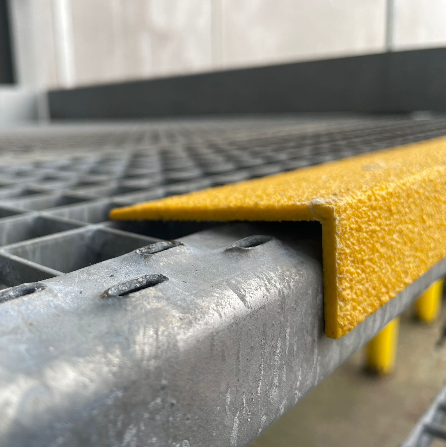 Close-up van een geel antislip trapprofiel bevestigd op de rand van een gegalvaniseerde stalen roostervloer, met op de achtergrond meer gele profielen