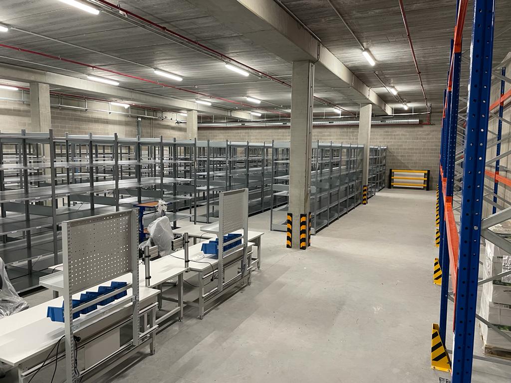 Overzicht van een nieuw ingericht ondergronds magazijn met grijze metalen stellingen, industriële werktafels met geperforeerde achterwanden en blauwe grijpbakken in een politiehoofdkwartier