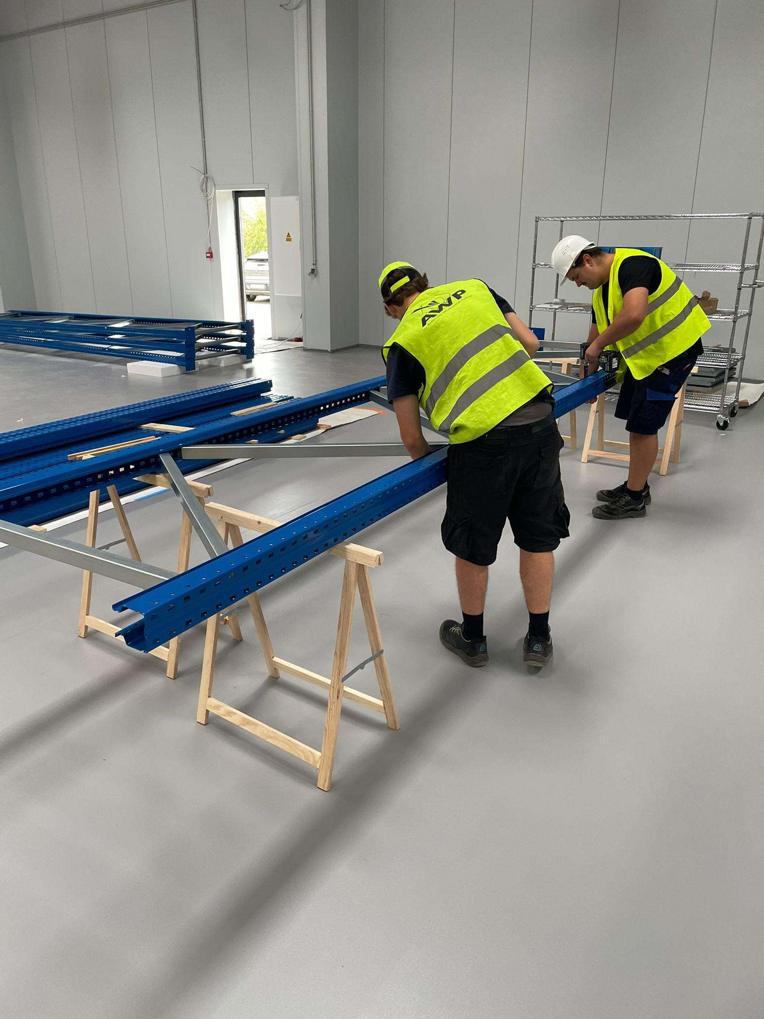 Twee AWP-medewerkers in fluogeel veiligheidsvest monteren blauwe palletrekliggers op houten schragen in een nieuwe magazijnhal