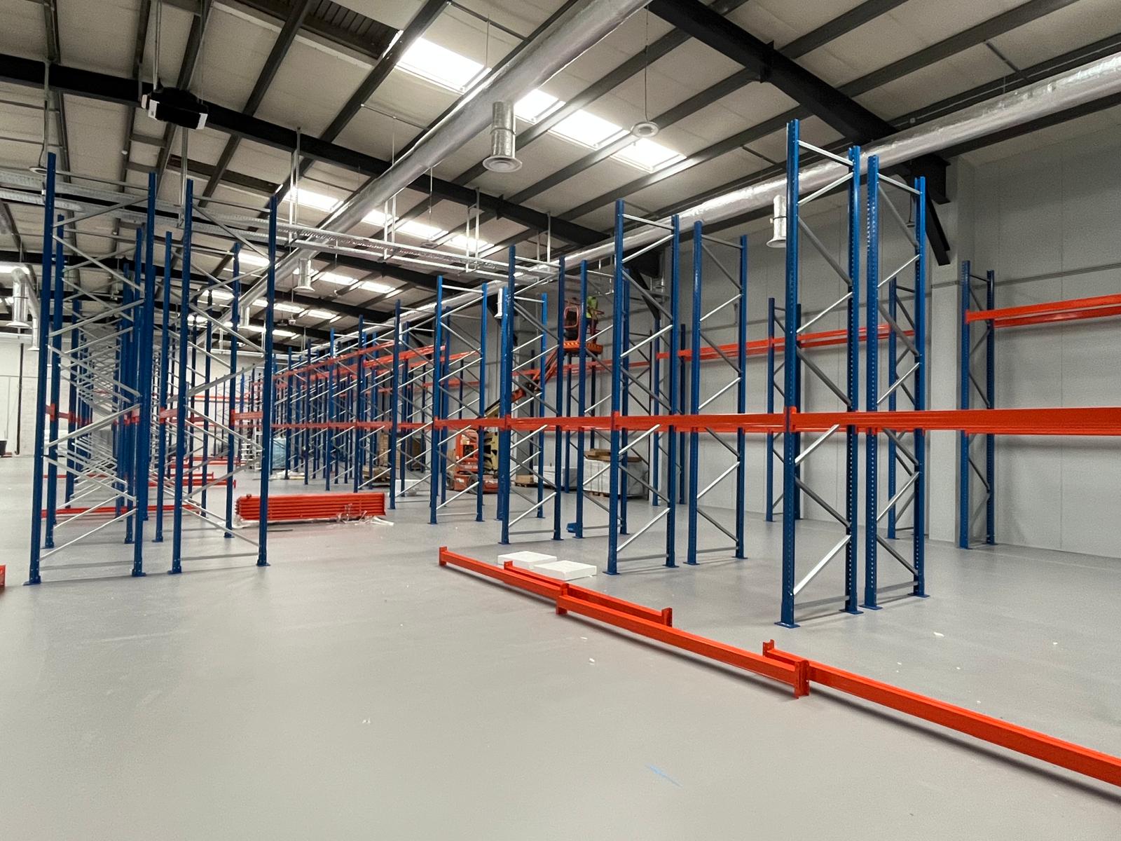 Overzicht van een grote magazijnhal met palletrekken in aanbouw, blauwe staanders en oranje liggers gedeeltelijk geplaatst, met oranje liggers op de vloer en een hoogwerker op de achtergrond