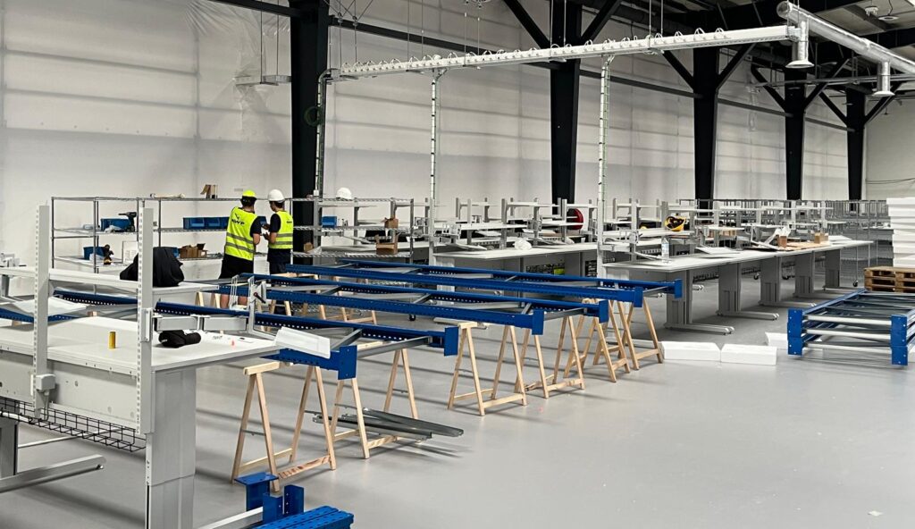 Twee AWP-medewerkers in fluogeel veiligheidsvest en helm monteren industriële werktafels in een nieuwe productiehal van een farmaceutisch bedrijf, met blauwe rolbaanonderdelen op schragen op de voorgrond