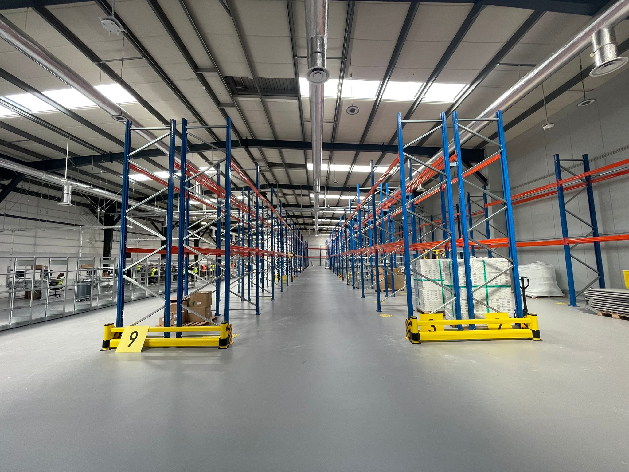 Breed gangpad tussen twee rijen hoog zwaar palletrek met blauwe staanders en oranje liggers in een nieuwe magazijnhal, met geel genummerde aanrijdbeveiligingen op de voorgrond