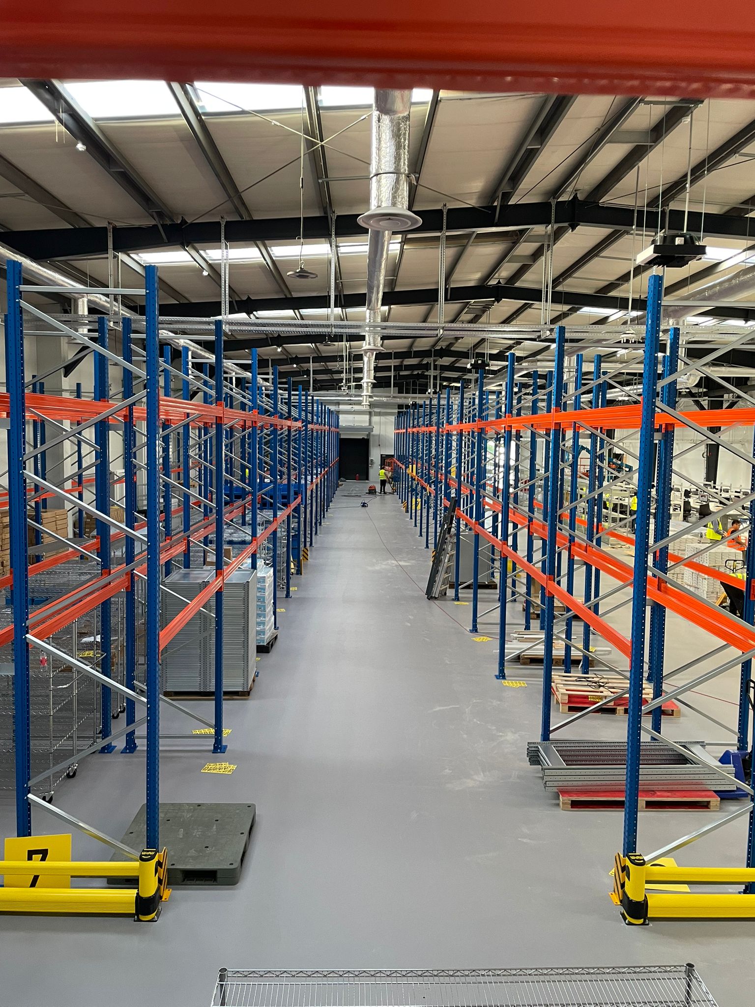 Bovenaanzicht vanop een hoog niveau van een breed gangpad tussen twee rijen zwaar palletrek met blauwe staanders en oranje liggers in een magazijnhal in inrichtingsfase, met gele aanrijdbeveiligingen op de vloer