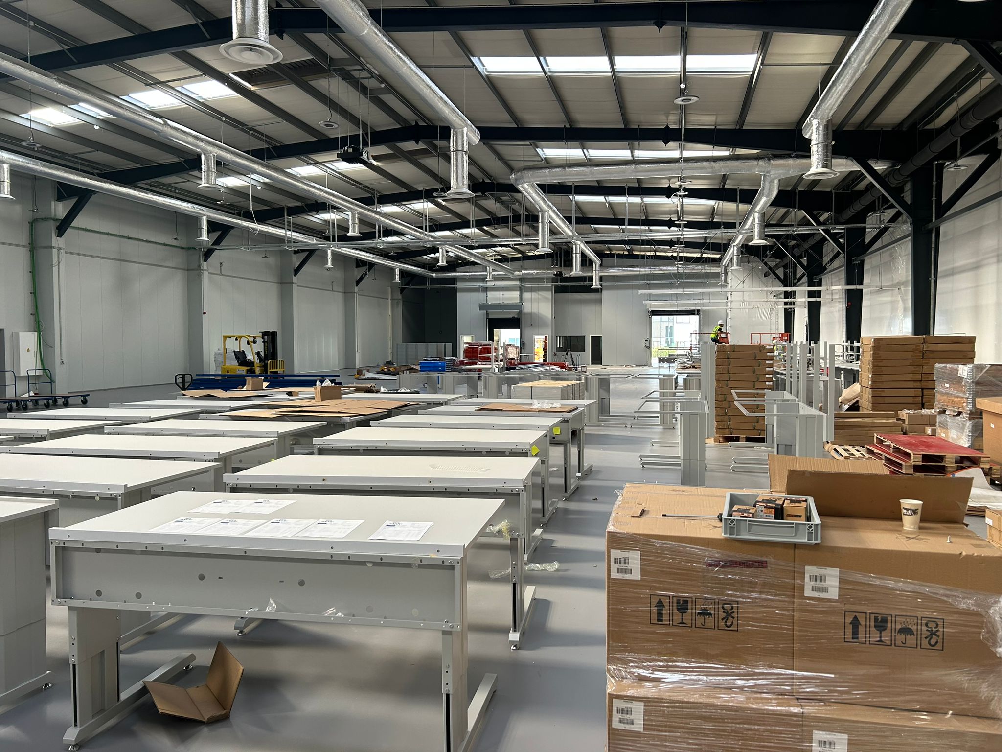 Meerdere rijen witte industriële werktafels in aanbouw in een grote productiehal, met pallets dozen en verpakkingsmateriaal op de voorgrond