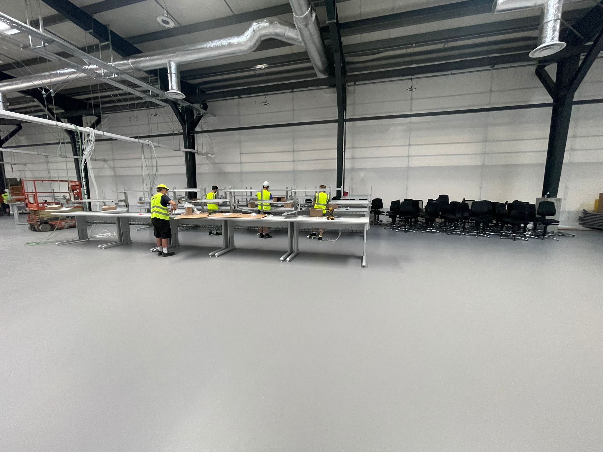 Vier AWP-medewerkers in veiligheidsvest monteren een rij industriële werktafels in een grote productiehal, met een groep zwarte bureaustoelen klaar voor gebruik op de achtergrond