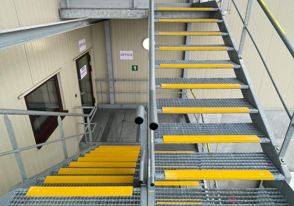 Bovenaanzicht van een verzinkte industriële buitentrap met gele antislip trapneuzen, toegang gevend tot een kantoorgebouw in een logistieke omgeving