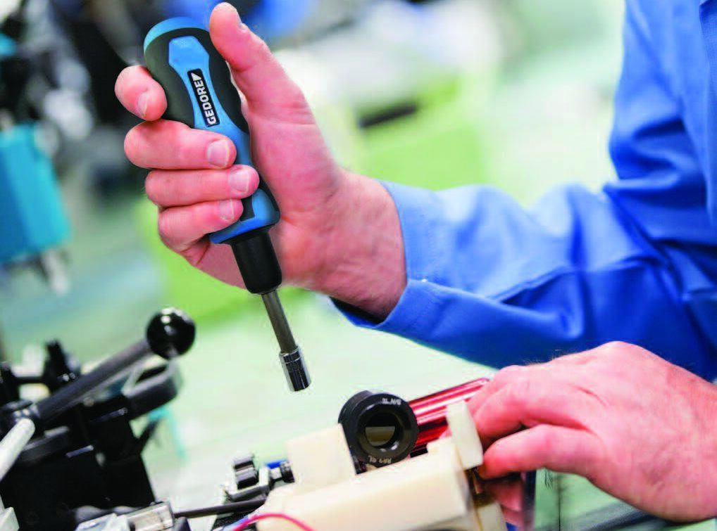 Een technicus in een blauwe werkjas gebruikt een blauw-zwarte Gedore schroevendraaier om een schroef vast te draaien in een precisie-apparaat of meetinstrument. Op de voorgrond zijn mechanische componenten zichtbaar, op de achtergrond een industriële werkomgeving. Un technicien en blouse bleue utilise un tournevis Gedore bleu et noir pour visser une vis dans un appareil de précision ou un instrument de mesure. Des composants mécaniques sont visibles au premier plan, avec un environnement de travail industriel en arrière-plan.