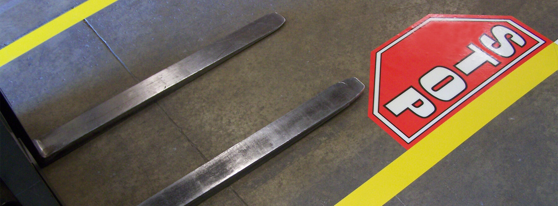 Vorken van een vorkheftruck die tot aan een gele vloermarkeringslijn reiken, met daarop een rood zeshoekig stopbordje als vloersticker op een betonnen magazijnvloer
