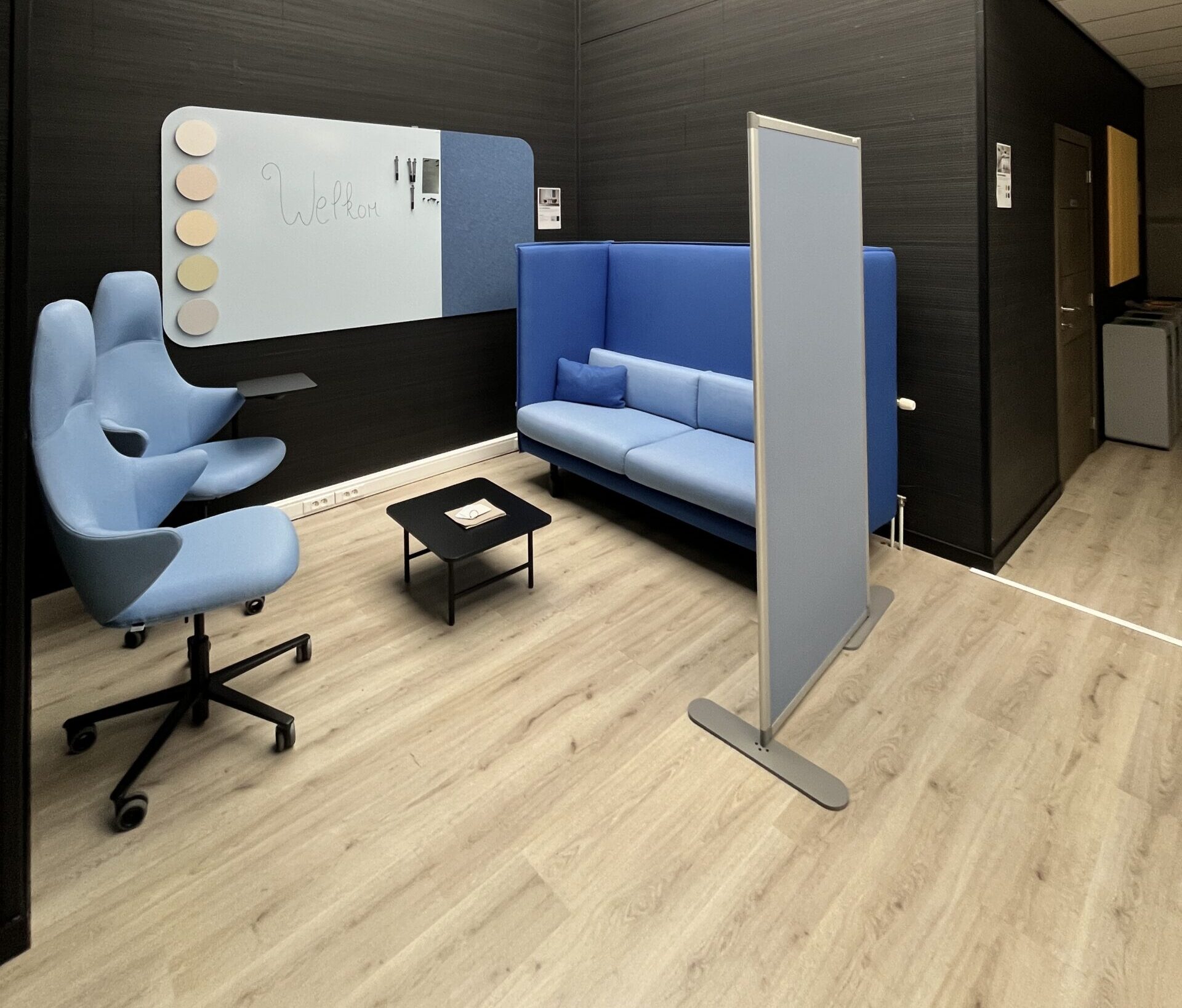 Lichte demoruimte, twee lichtblauwe draaifauteuils, een blauwe akoestische loungebank, een combinatie whiteboard-prikbord en een staande scheidingswand