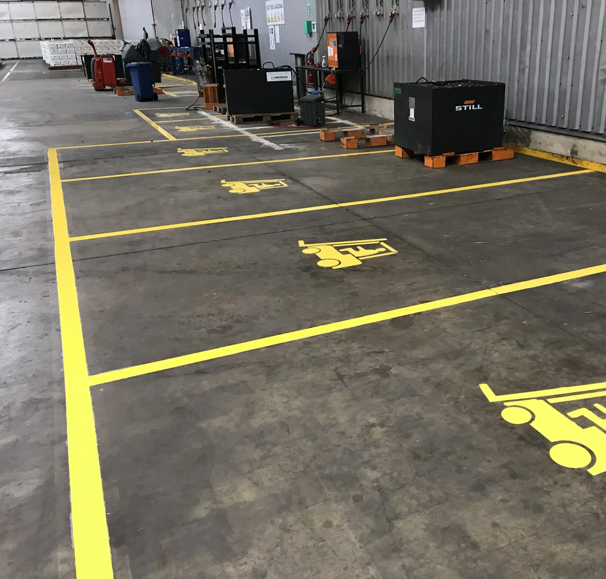 Betonnen magazijnvloer met gele lijnen en herhaalde vorkheftruckpictogrammen die parkeerplaatsen voor heftrucks afbakenen / Sol d'entrepôt en béton avec des lignes jaunes et des pictogrammes répétés de chariot élévateur délimitant des emplacements de stationnement