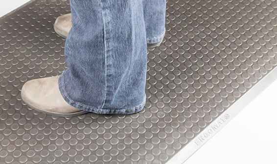 Onderbenen van persoon in spijkerbroek en lichte schoenen staand op een grijze Ergomat Complete Smooth mat met fijn noppenpatroon en zilverkleurige rand