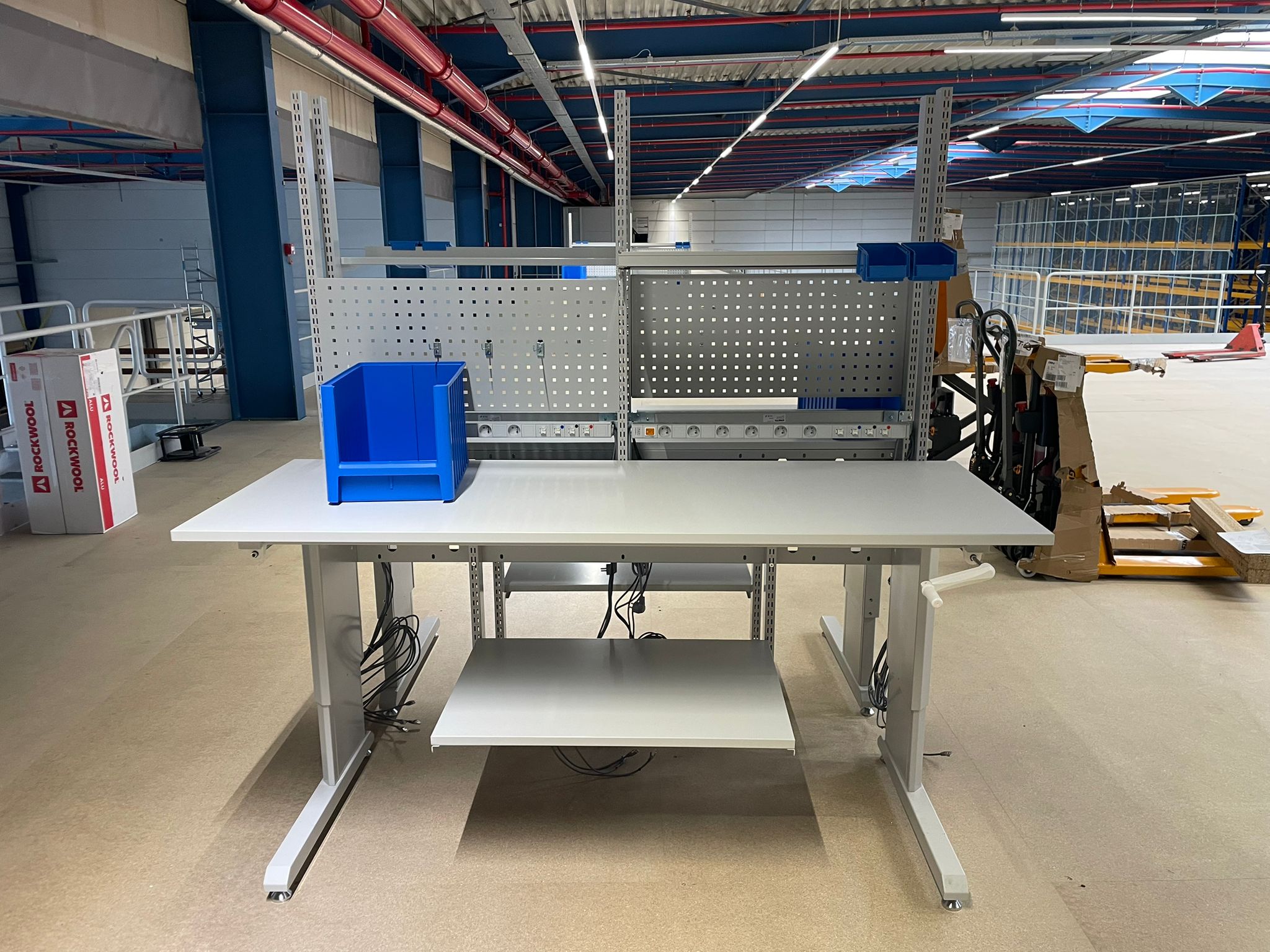 In hoogte verstelbare werktafel met handzwengel, prikbord en magazijnbak als onderdeel van de ergonomische werkposten in het magazijn van Pro@Work
