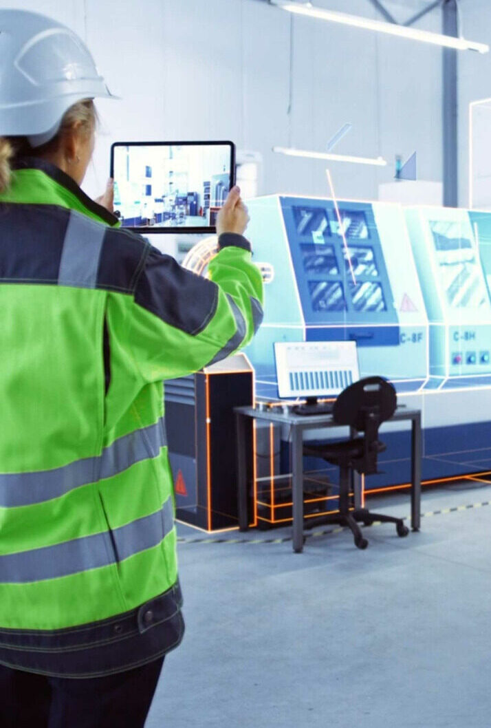 Medewerker in groene veiligheidsvest en witte helm gebruikt een tablet in een industriële productiehal met CNC-machines op de achtergrond, waarbij digitale AR-overlays zichtbaar zijn.