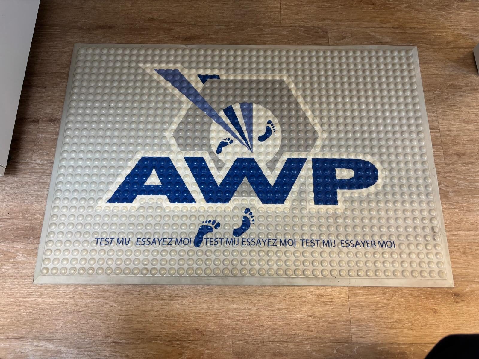 antivermoeidheidsmat met blauwe AWP-logobedrukking, voetafdrukken en de teksten "Test mij" en "Essayez moi", liggend op een houten vloer