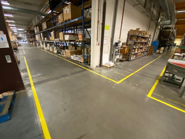 Gele vloermarkering die rijroutes en werkzones afbakent in de productiehal van Brepols