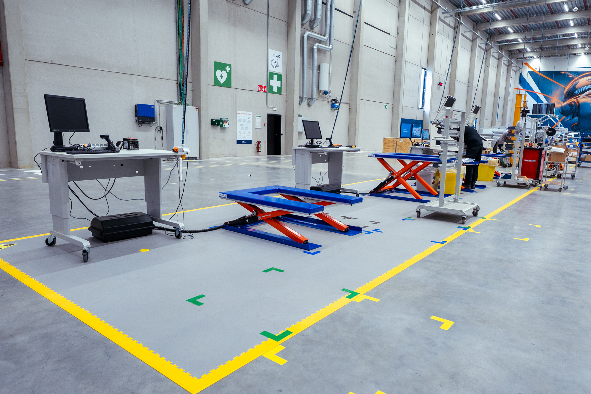 AWP Productiehal met in hoogte verstelbare werktafels, blauw-oranje schaarheftafels en gele vloermarkering rond de werkposten. - AWP Hall de production avec postes de travail réglables en hauteur, tables élévatrices à ciseaux et marquage au sol jaune.
